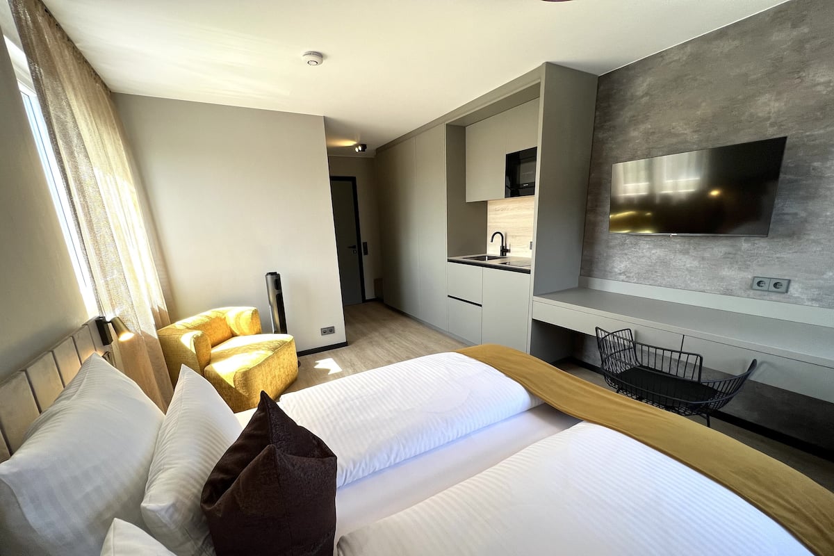 A modern hotel room with a large bed, a desk, a chair, a TV, and a kitchenette.
