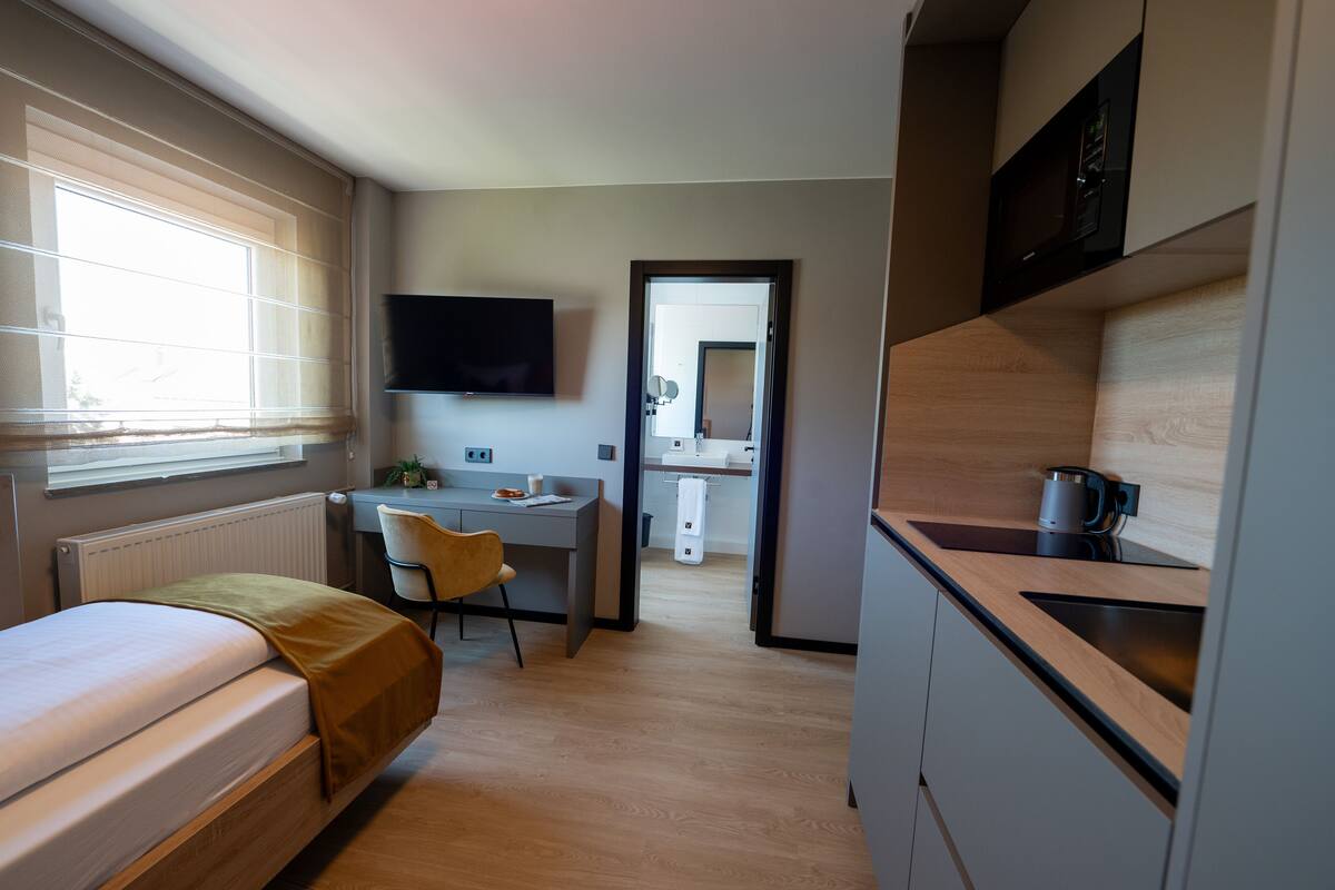 A compact hotel room with a bed, a desk, a chair, a TV, a kitchenette, and a bathroom visible through an open door.
