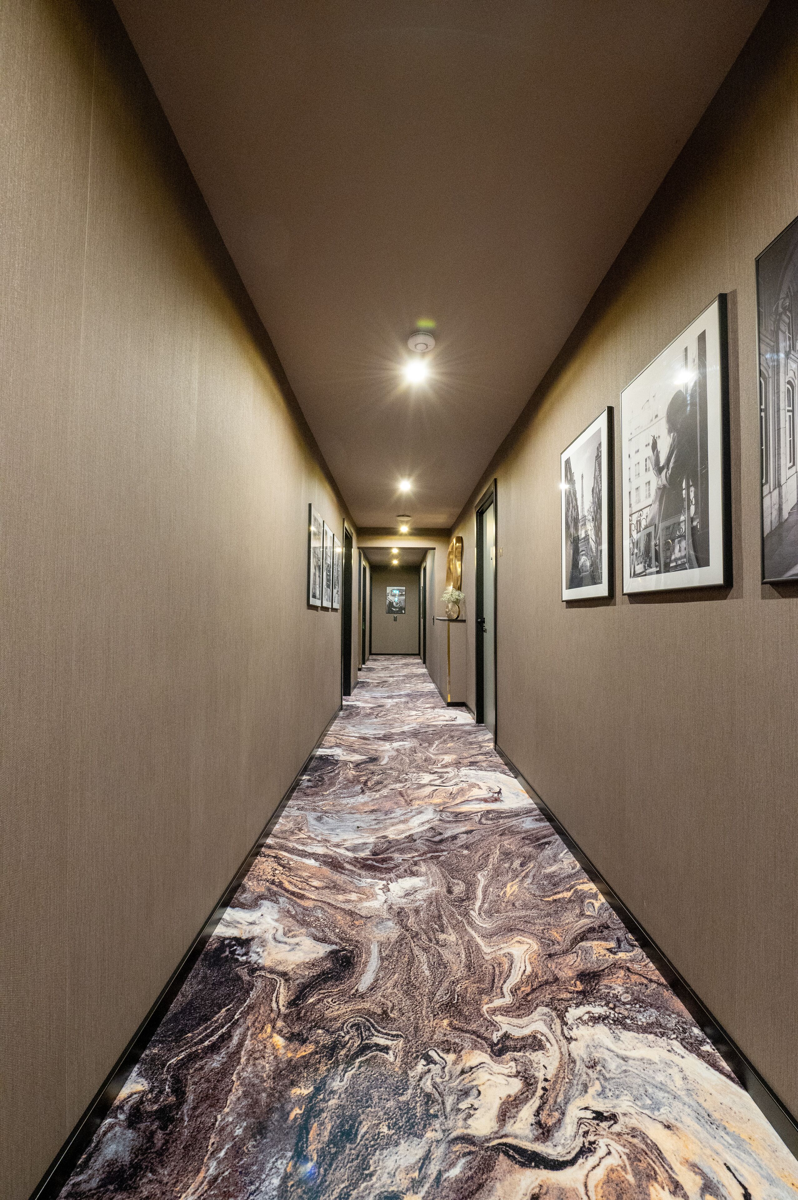 A hotel hallway with a marble-patterned carpet, framed pictures on the walls, and a ceiling light.