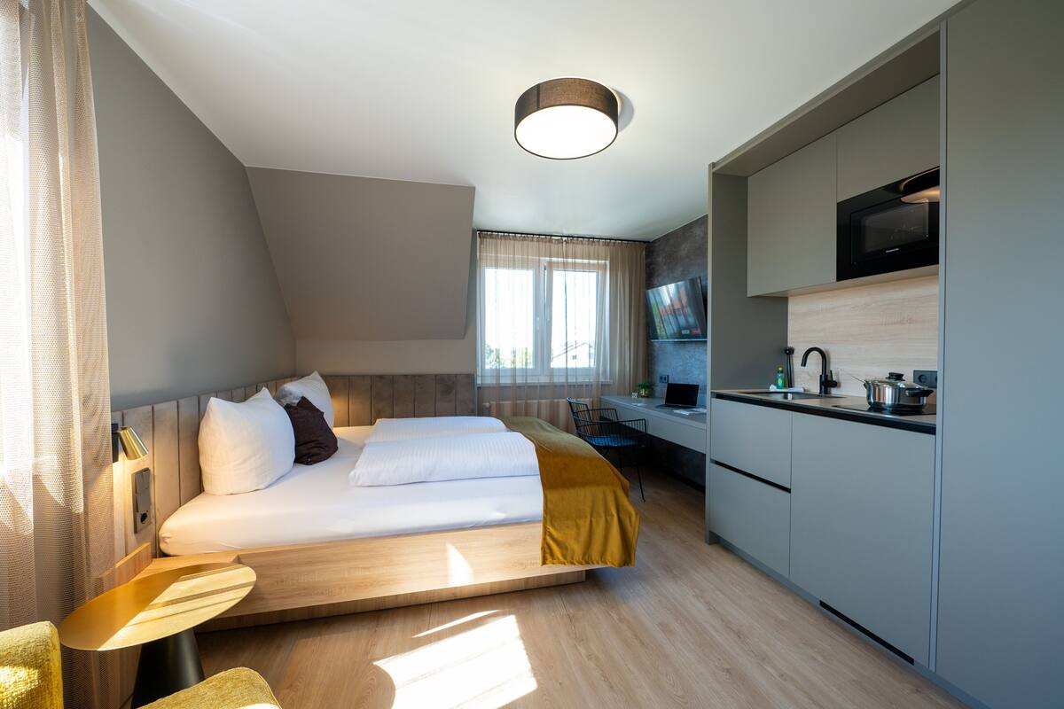 A modern hotel room with a bed, a desk, a kitchenette, and a TV.