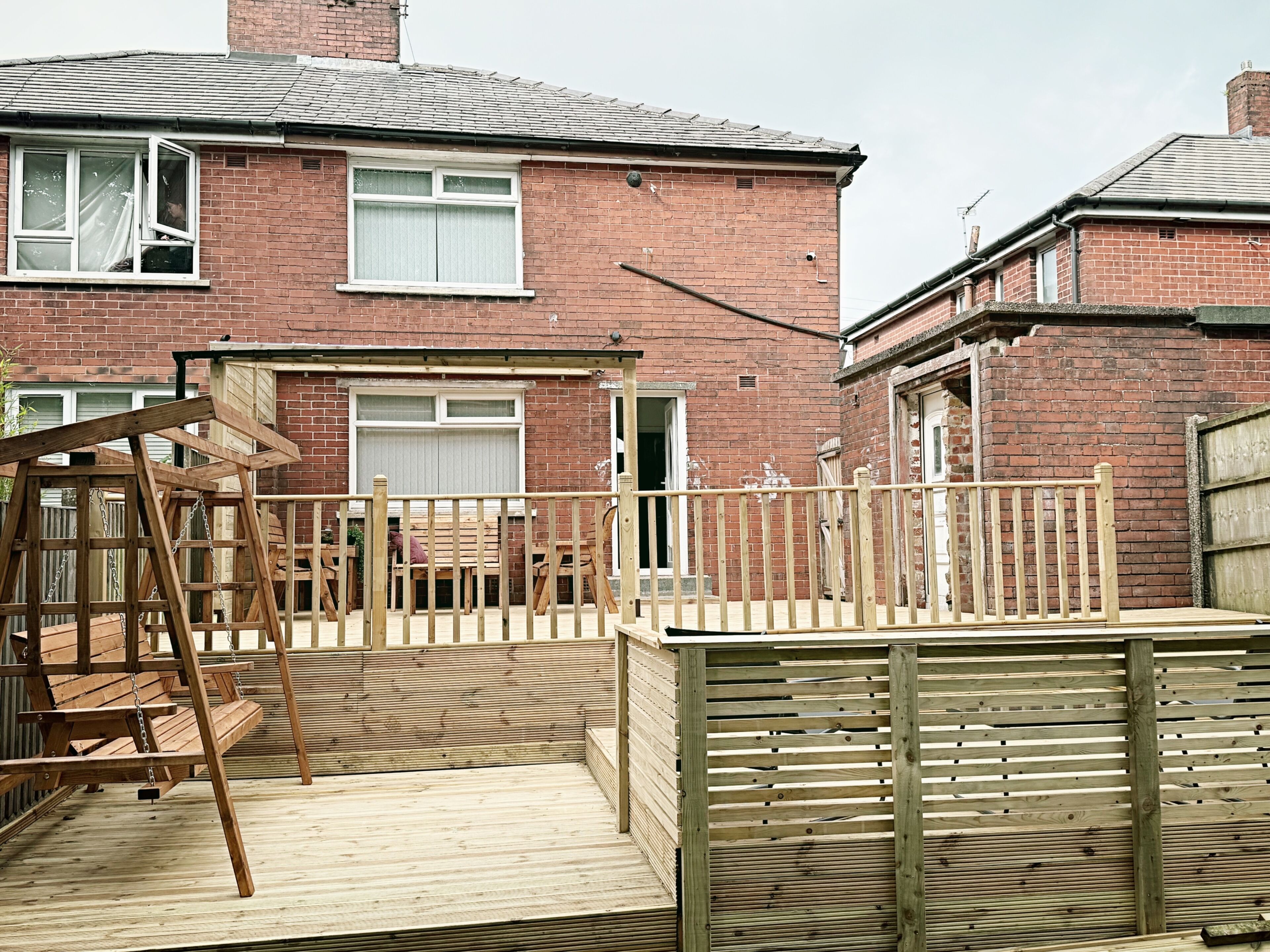 A residential backyard with a wooden deck, a swing set, and a brick house.