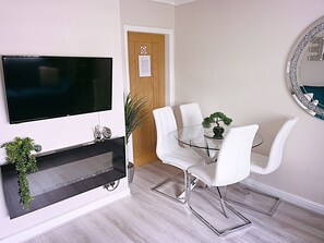 A modern dining area with a glass table, white chairs, a TV mounted on the wall, and a small bonsai tree on the table. - The Serene Stays Sleeps 6 With Garden (Oldham)