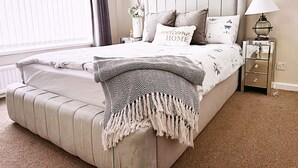 A neatly made bed with a 'welcome home' pillow, a bedside lamp, and a nightstand with a mirror. - The Serene Stays Sleeps 6 With Garden (Oldham)