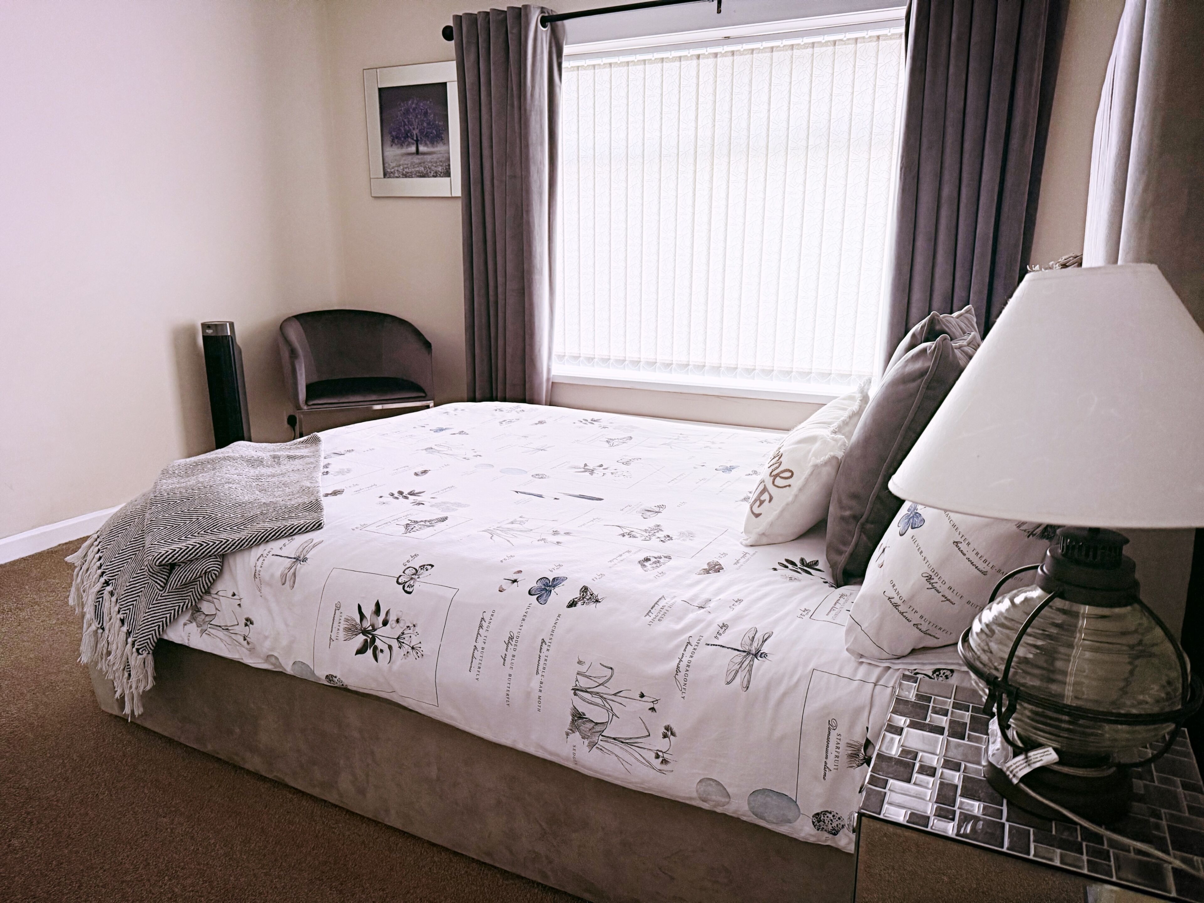 A neatly made bed with a floral-patterned bedspread, a bedside lamp, and a chair in a room with a window and curtains.