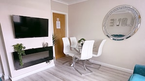A modern living room with a flat-screen TV mounted on the wall, a dining table with white chairs, a small potted plant, and a decorative mirror with the word "LOVE" on the wall. - The Serene Stays Sleeps 6 With Garden (Oldham)