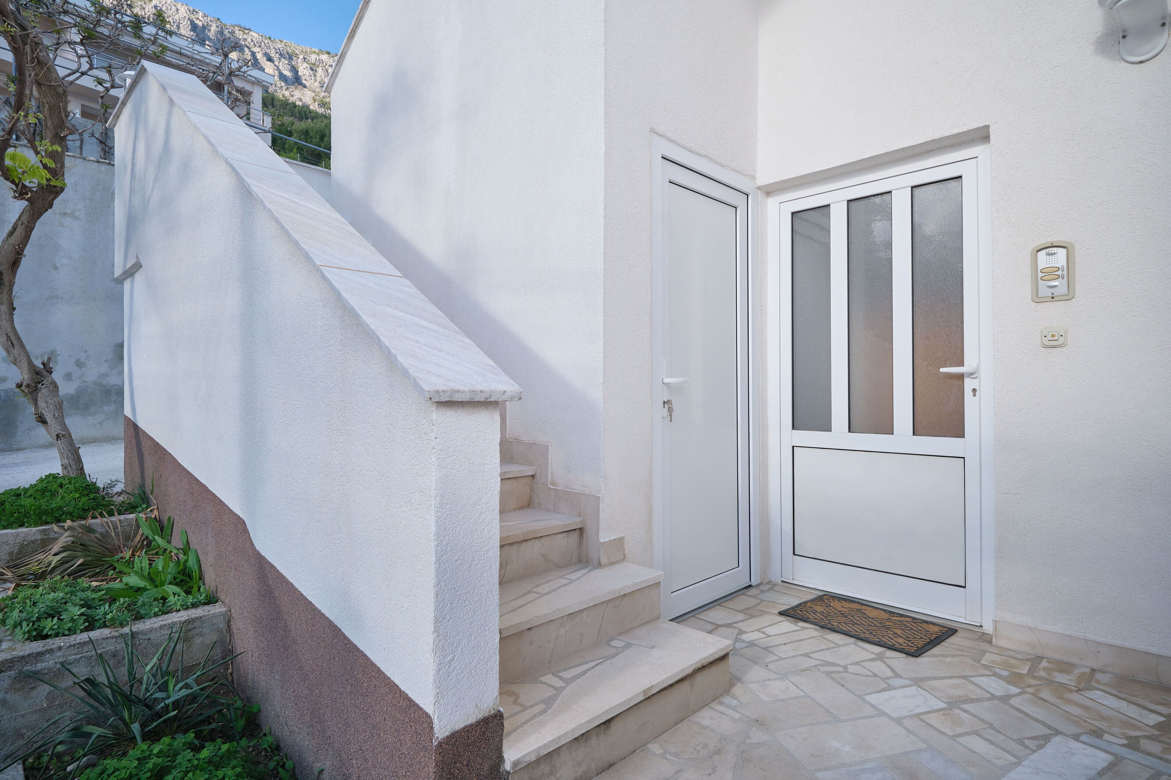A white exterior with a set of stone steps leading to a door, a small garden area with plants, and a stone-tiled entrance.