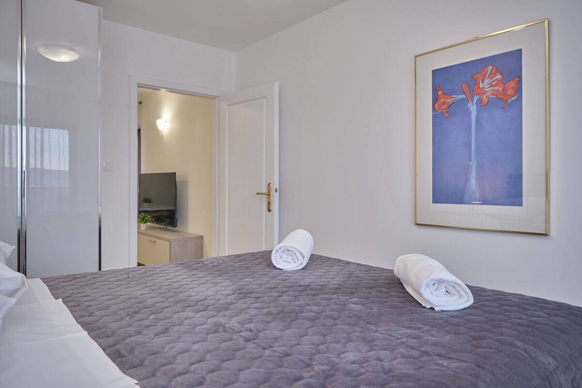 Una cama bien hecha con toallas enrolladas, un cuadro enmarcado en la pared y una puerta que da a otra habitación.
