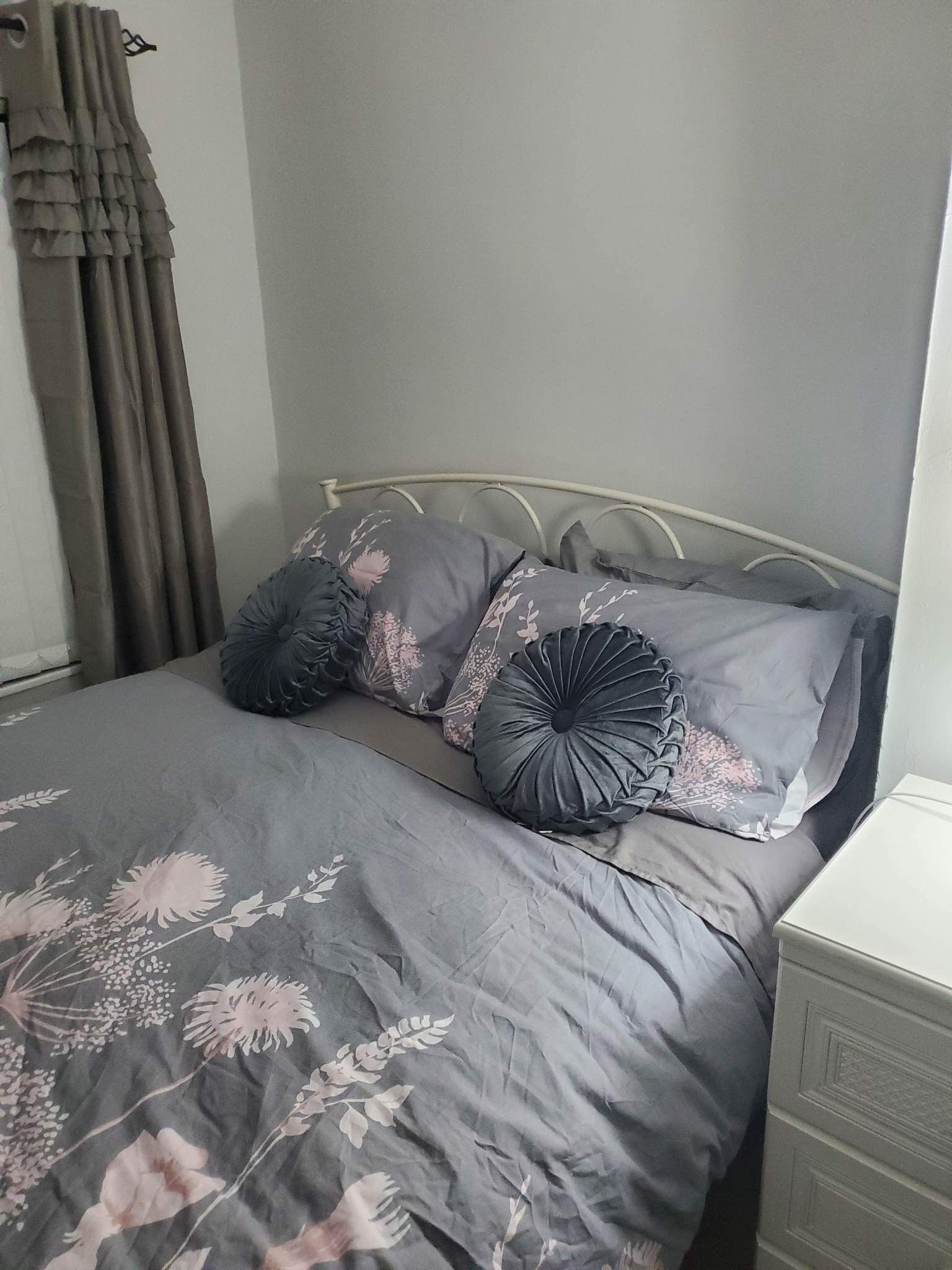 Un letto con copripiumino floreale grigio, due cuscini grigi con motivi floreali e una testiera in metallo bianco.