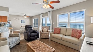 A living room with a ceiling fan, a sofa, a chair, a coffee table, and a view of the ocean through large windows. - IBR 5006: Ocean Star - Catch the Breeze and Feel the Peace! (Fort Walton Beach)