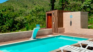 A swimming pool with a wooden deck and lounge chairs, surrounded by greenery and a mountain in the background.
