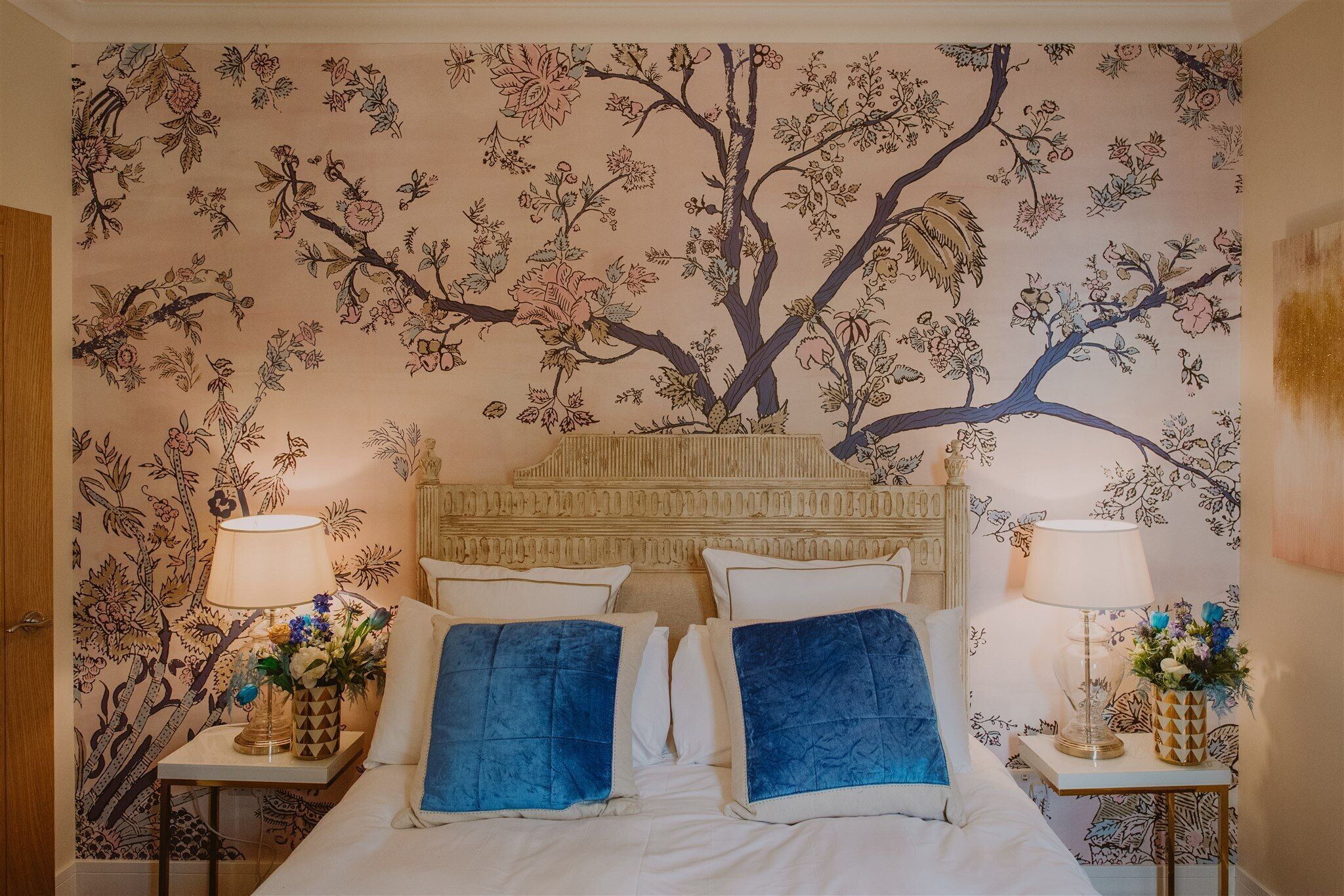 A bedroom with a floral wallpaper, a wooden headboard, two bedside tables with lamps, and blue pillows.
