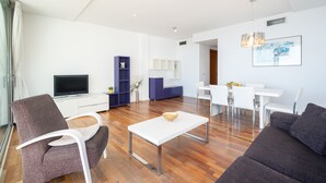 Un salon moderne avec un téléviseur à écran plat, une table basse blanche et un canapé. On y trouve également un coin repas avec une table et des chaises blanches. La pièce est dotée d’un plancher en bois et de murs blancs.
