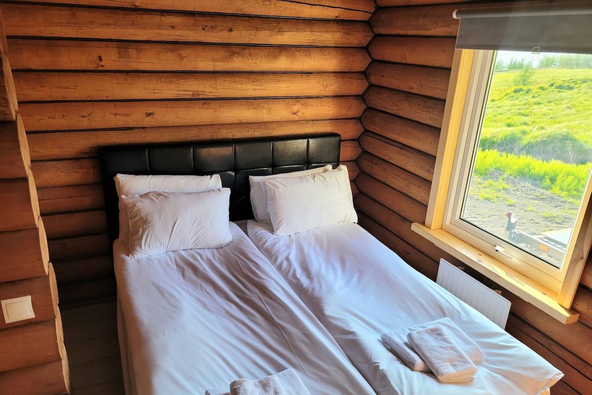 Habitación de cabaña de madera con una cama, dos almohadas y ventana con vistas a un paisaje cubierto de hierba.