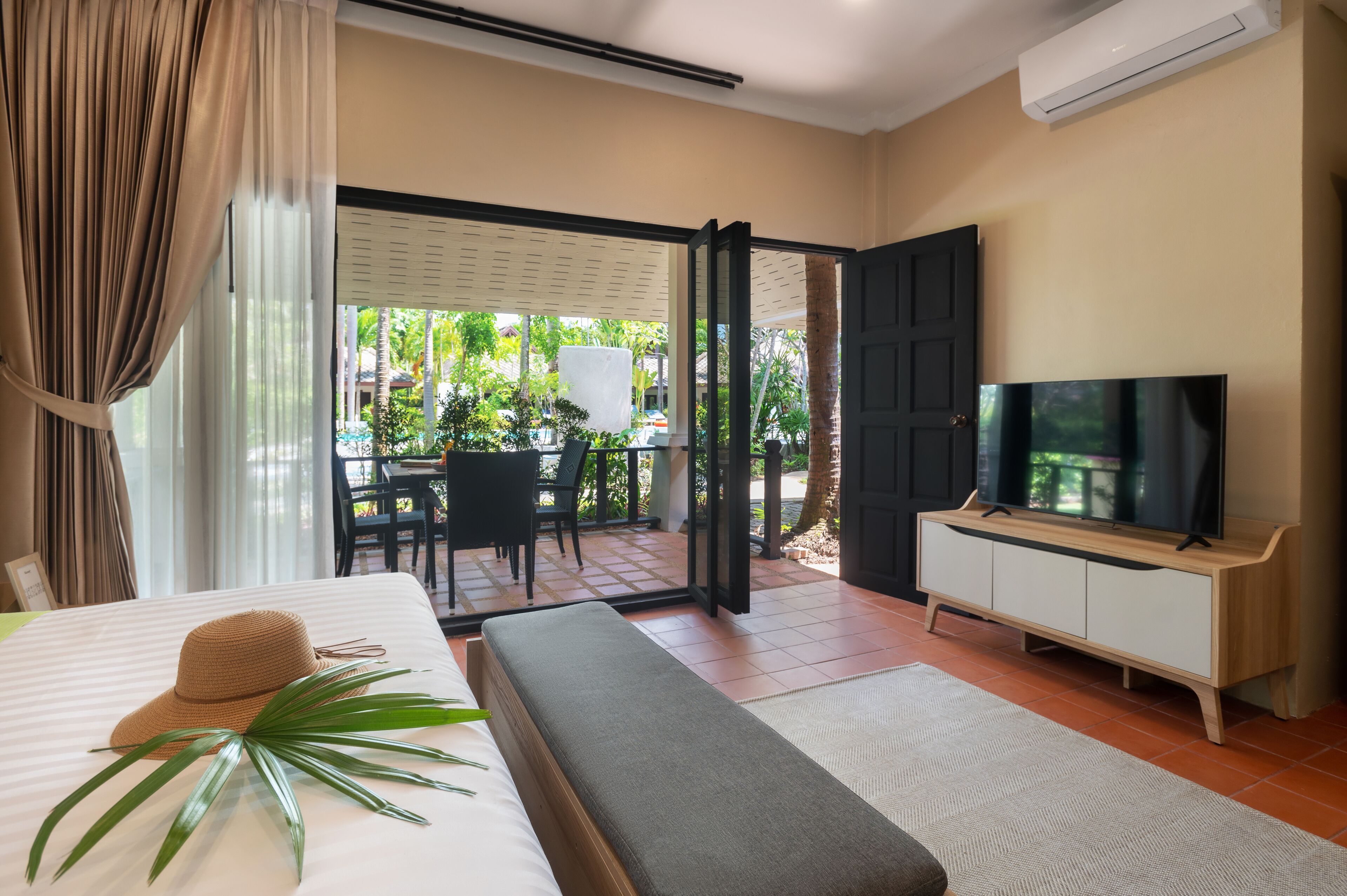 Habitación de hotel con cama, televisor, banca, planta y vista al área exterior.