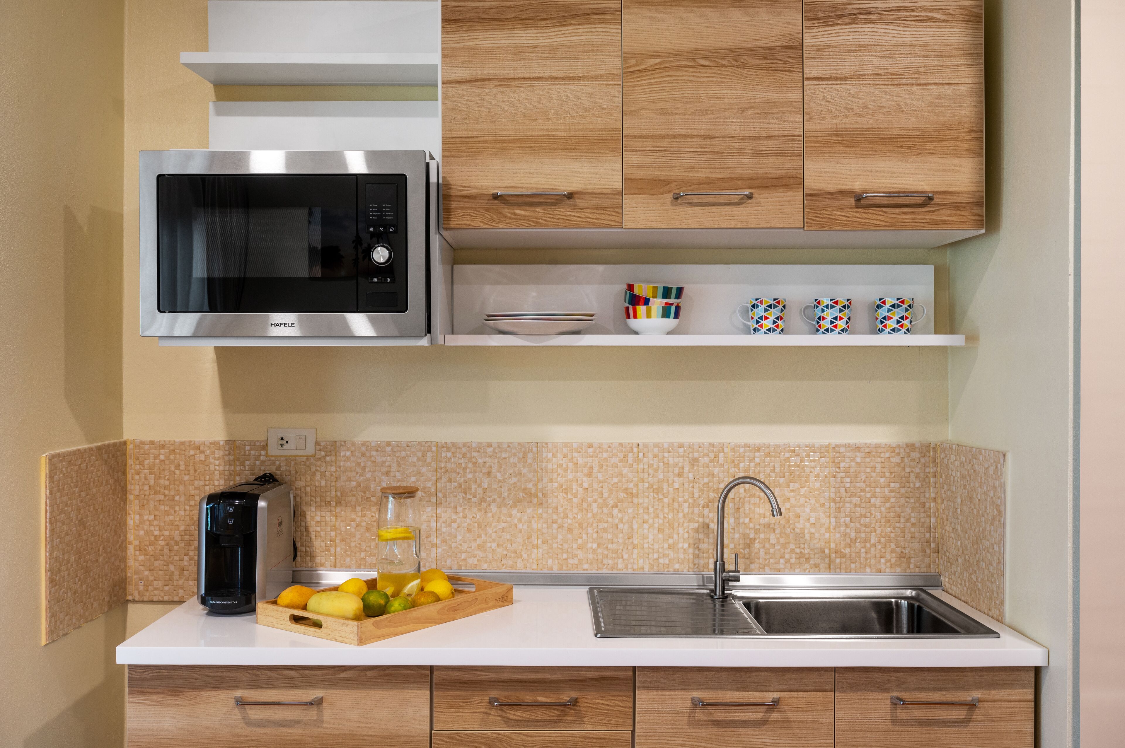 A modern kitchen with a built-in microwave, a coffee maker, a fruit bowl, and a stainless steel sink.