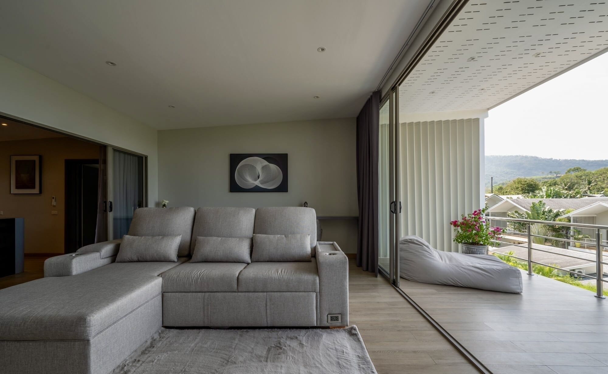 A modern living room with a grey sectional sofa, a large window with a city view, and a balcony with plants.