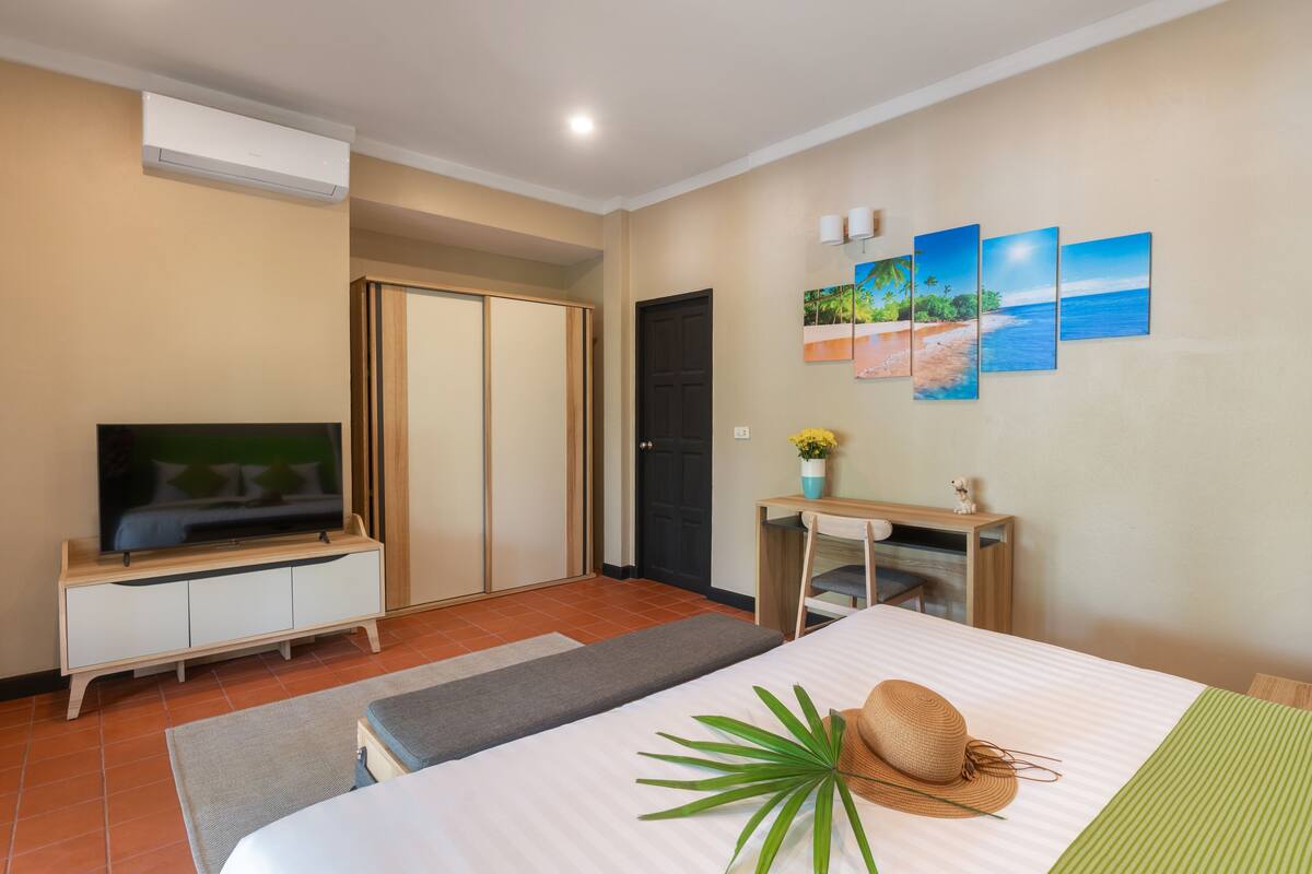 A hotel room with a bed, a TV, a wardrobe, a desk, and a wall with beach paintings.
