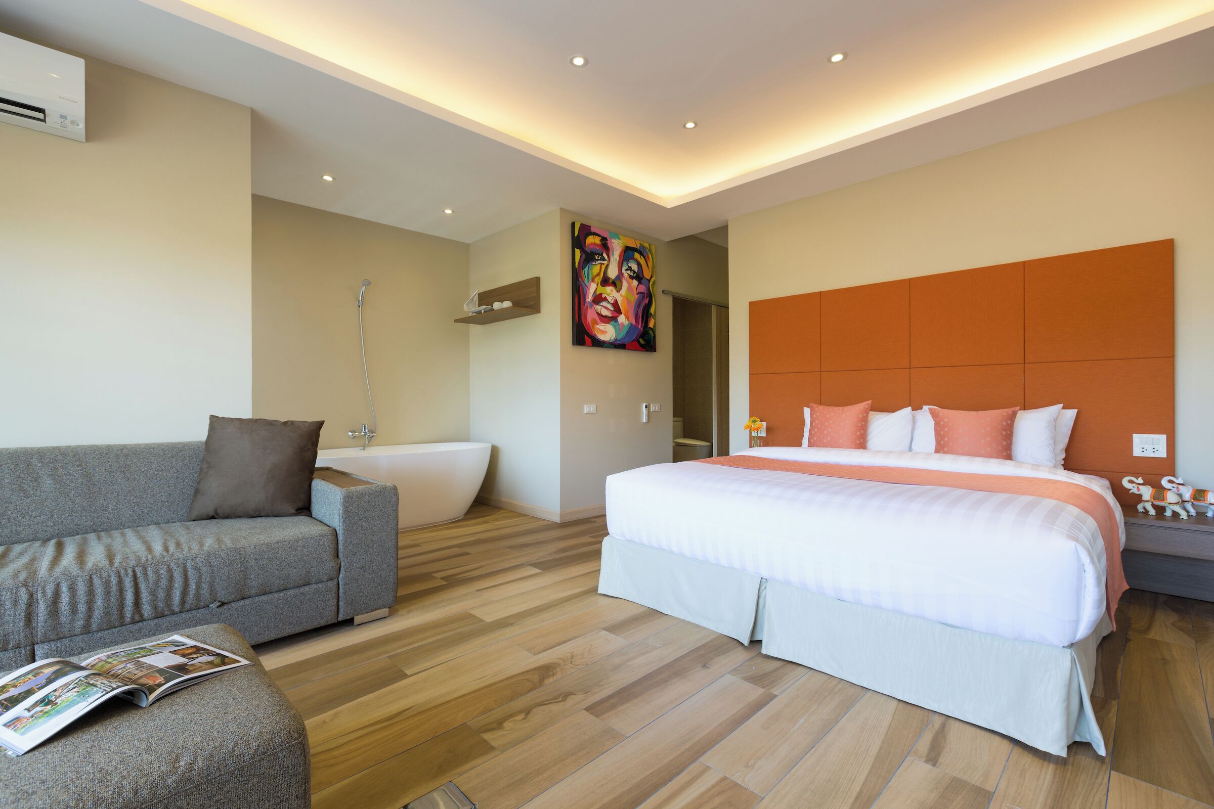 Une chambre d’hôtel moderne avec un canapé gris, un grand lit avec une literie orange, une baignoire blanche et un tableau coloré au mur.