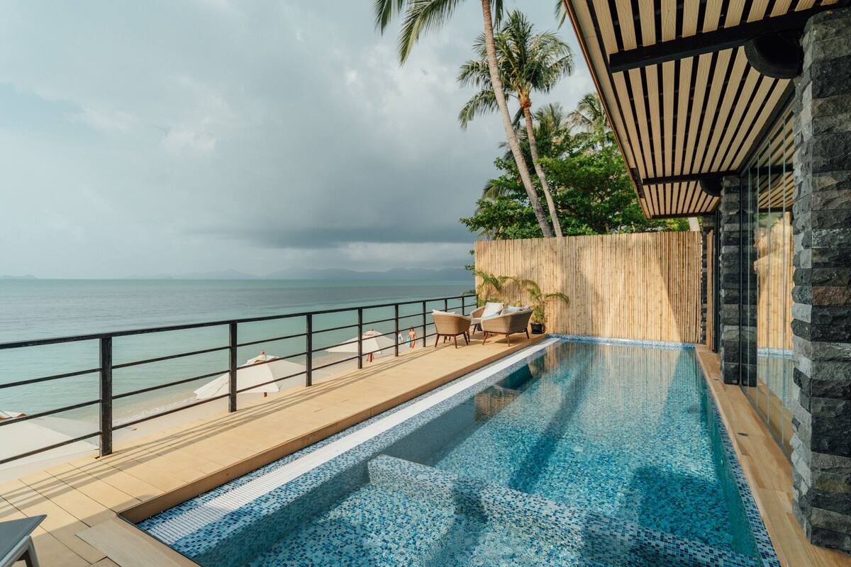 A pool with a view of the ocean, wooden deck chairs, and a stone wall.