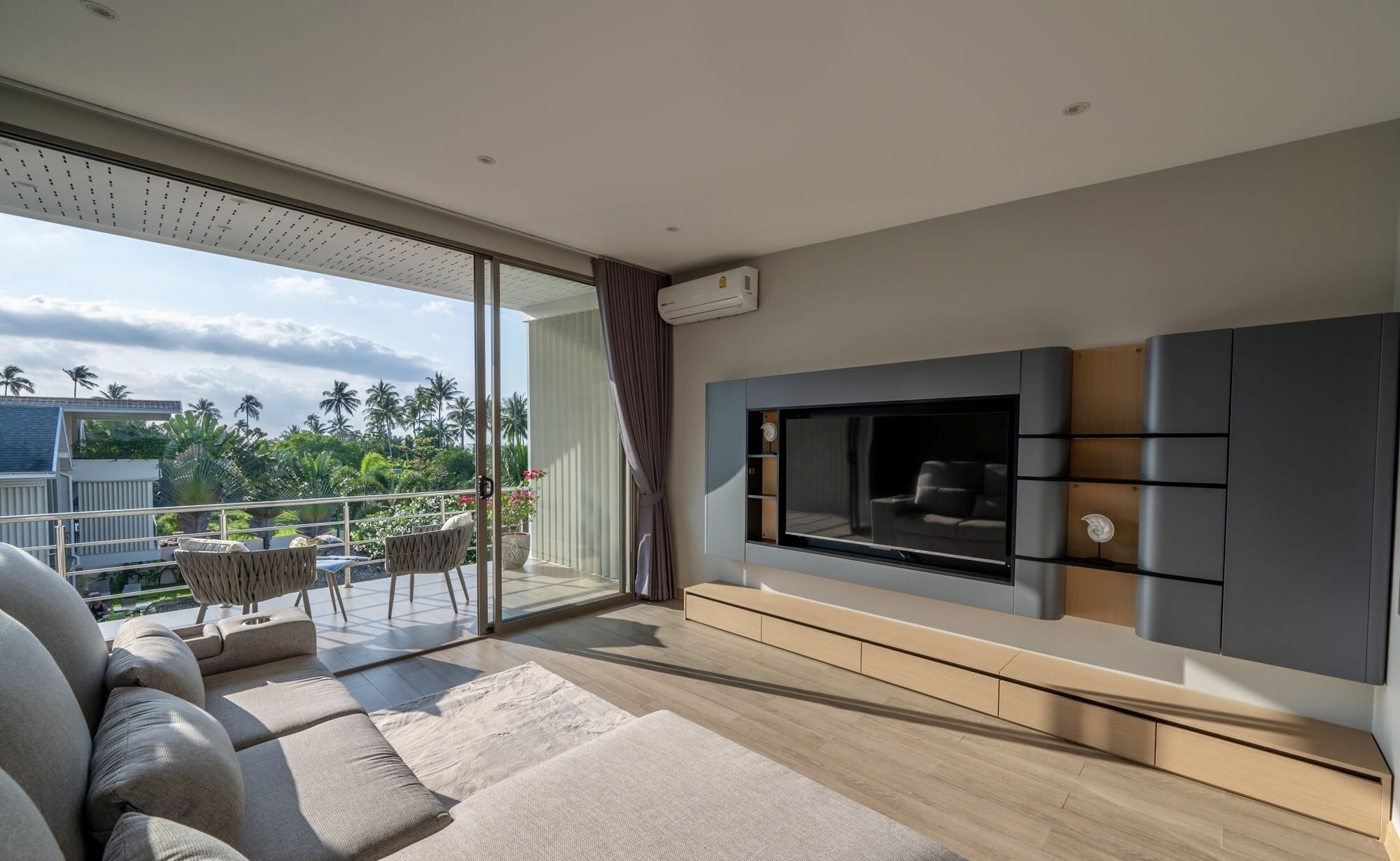 A modern living room with a large TV, comfortable seating, and a balcony with a view of palm trees.