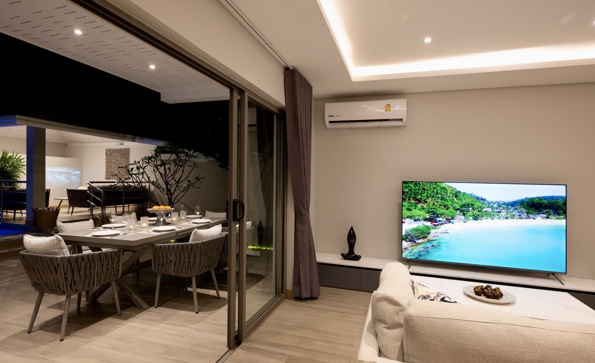A modern living room with a dining area, a flat-screen TV, and a view of a beach.