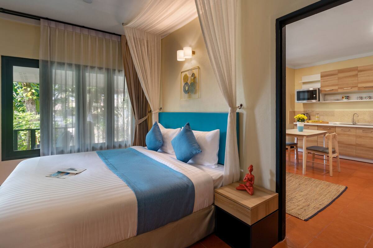 A hotel room with a bed, a nightstand, a window with curtains, a view of the outside, and a kitchen area visible through an open door.