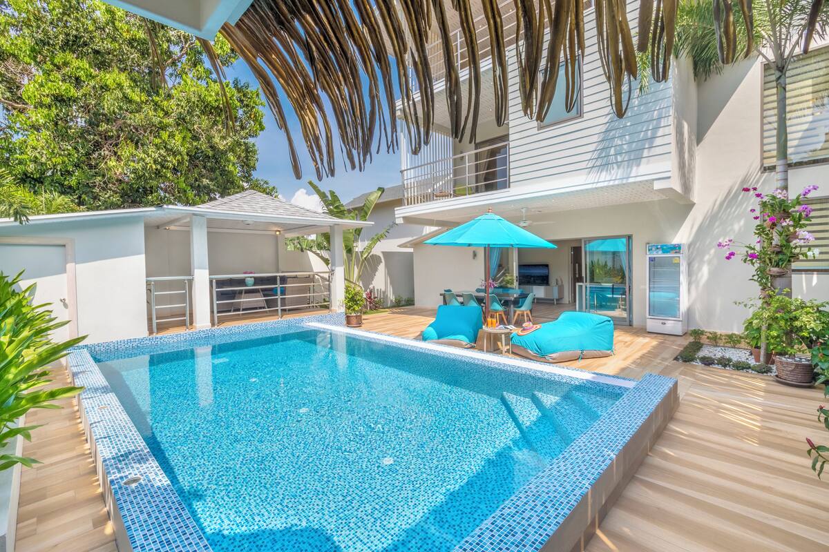 A swimming pool with a tiled edge, surrounded by wooden decking and outdoor furniture.