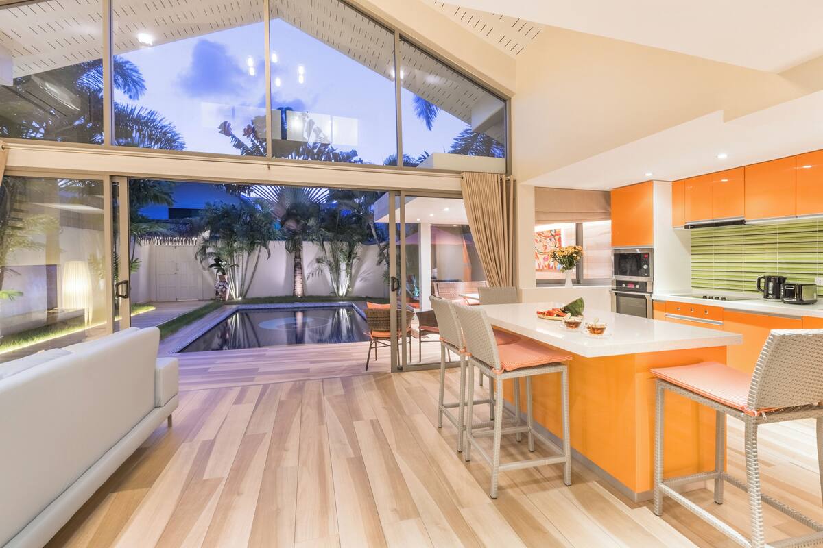 A modern kitchen with a large island, a dining area with chairs, and a view of a swimming pool and greenery.