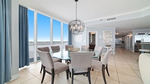 A dining area with a glass table and six upholstered chairs, large windows with blue curtains, and a view of the sky. - Oceania 5, New Owner, Impeccable Beachfront Condo!! (Gulf Shores)