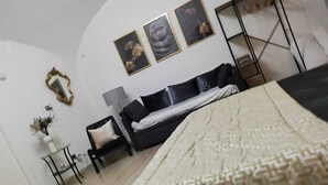 Un dormitorio con cama, sofá negro, una mesita de noche con lámpara y espejo en la pared.