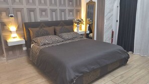 Un dormitorio con una cama grande, almohadas estampadas, una mesita de noche con lámpara, un espejo y una puerta con cortinas.