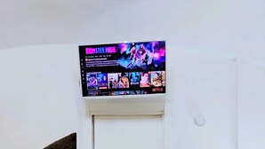 Una habitación con una puerta blanca, un televisor instalado mostrando un programa, una mesita con superficie de vidrio y un estante con varios objetos.