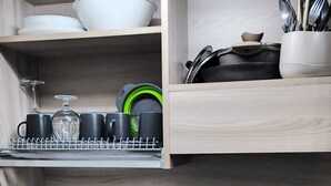 Una cocina con encimera negra, un lavamanos blanco y armarios de madera. Hay estantes con vajilla, vasos y una olla.