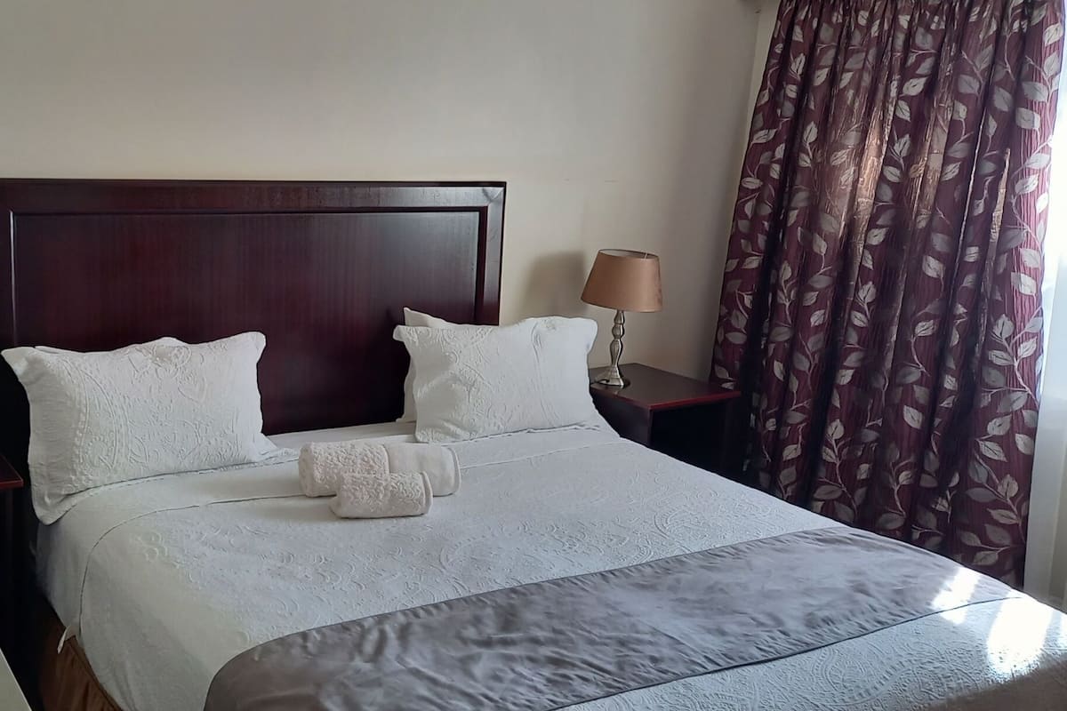 Een net opgemaakt bed met een houten hoofdbord, twee bedlampen en een raam met gordijnen.