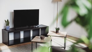 A modern living room with a flat-screen TV on a black TV stand, a small wooden coffee table, two potted plants, and a floor lamp.