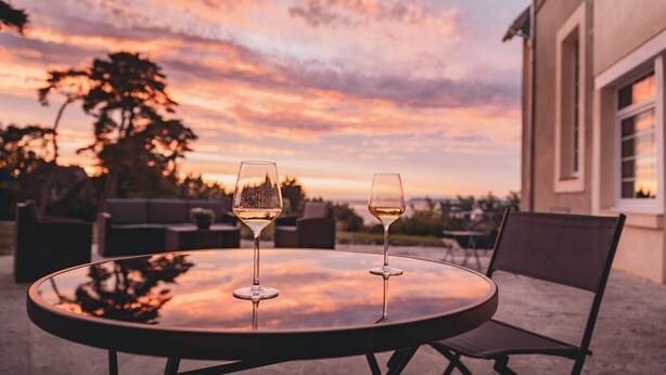 A round glass table with two wine glasses, a chair, and a view of a sunset over a landscape with trees and a building.