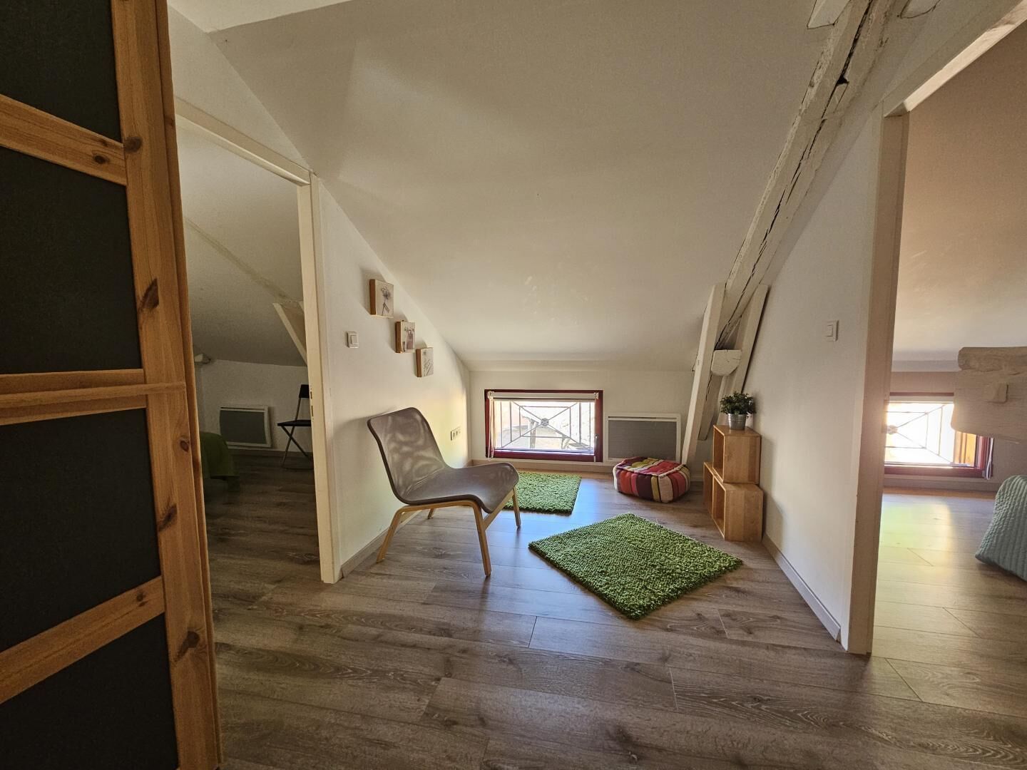 Een loft-achtige kamer met een hoge plafond, houten vloer, een groen vloerkleed, een stoel en een raam met uitzicht.