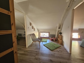 Une pièce de style « loft », avec un plafond haut, un plancher en bois, un tapis vert, une chaise et une fenêtre offrant une vue dégagée.