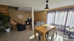 A living room with a wooden dining table, a sofa, a TV, and a staircase leading to an upper level. - Gîte du Séchoir - Bouresse (BOURESSE)