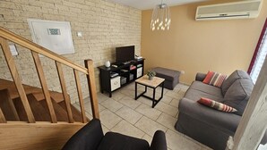 A living room with a staircase, a flat-screen TV, a wooden coffee table, a grey sofa, and a wall-mounted air conditioner. - Gîte du Séchoir - Bouresse (BOURESSE)