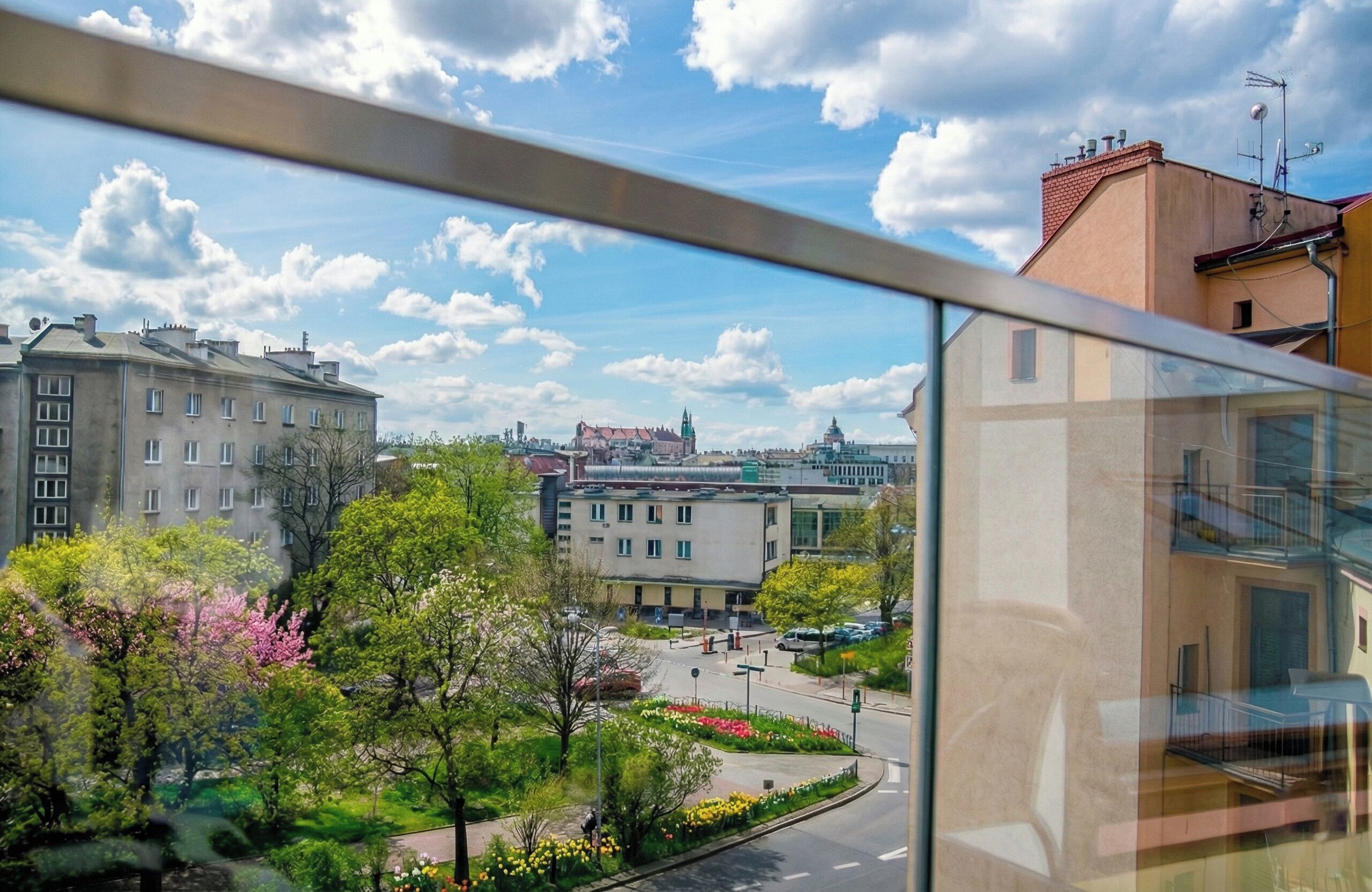 En utsikt från ett fönster med utsikt över en stadsbild med byggnader, en väg och en park med träd och blommor.