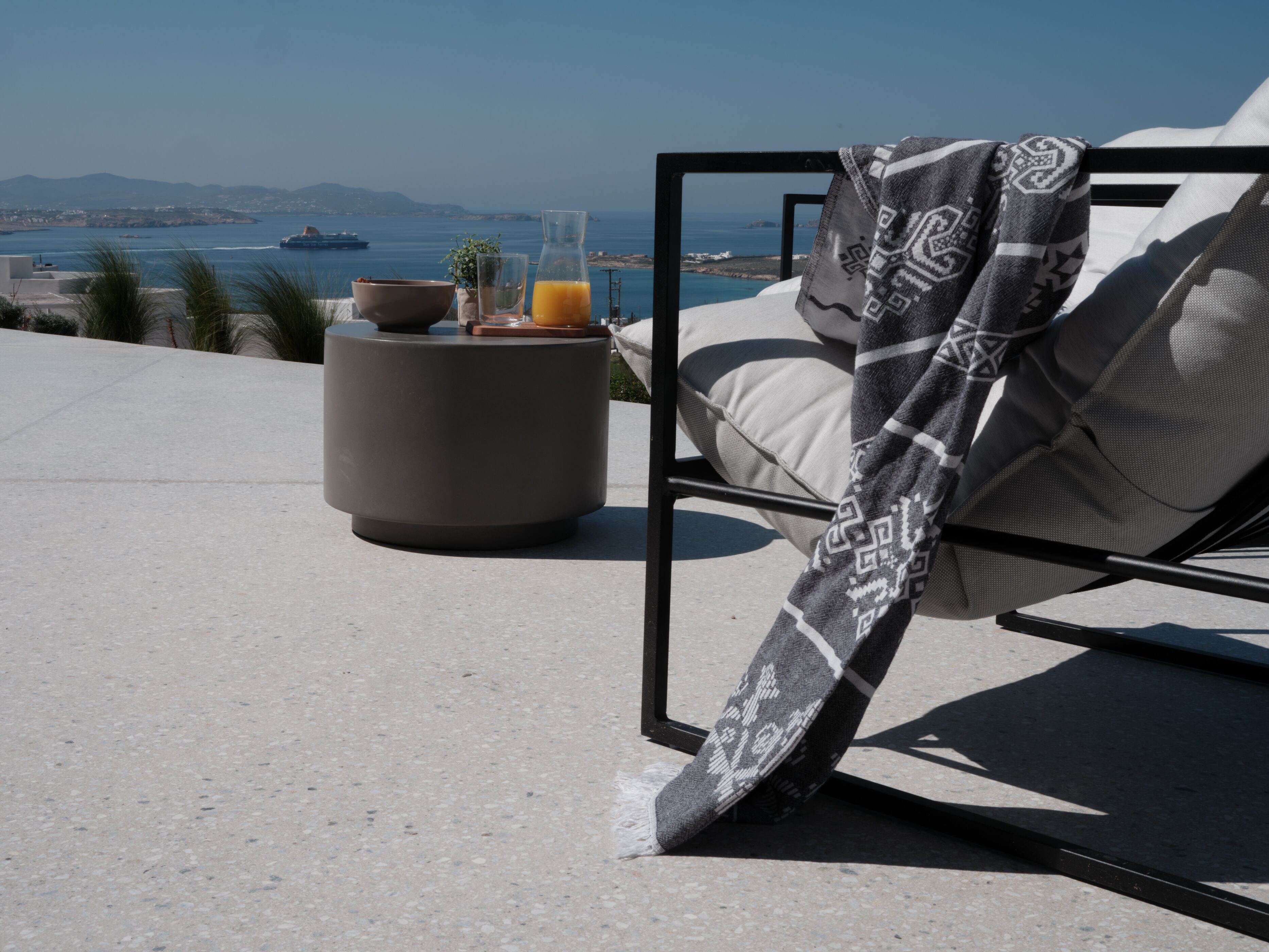 A lounge chair with a patterned throw, a glass of orange juice, and a bowl on a round table with a view of the sea and distant land.