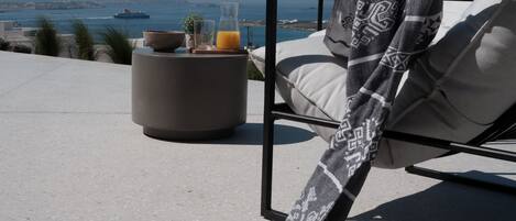 A lounge chair with a patterned throw, a glass of orange juice, and a bowl on a round table with a view of the sea and distant land.