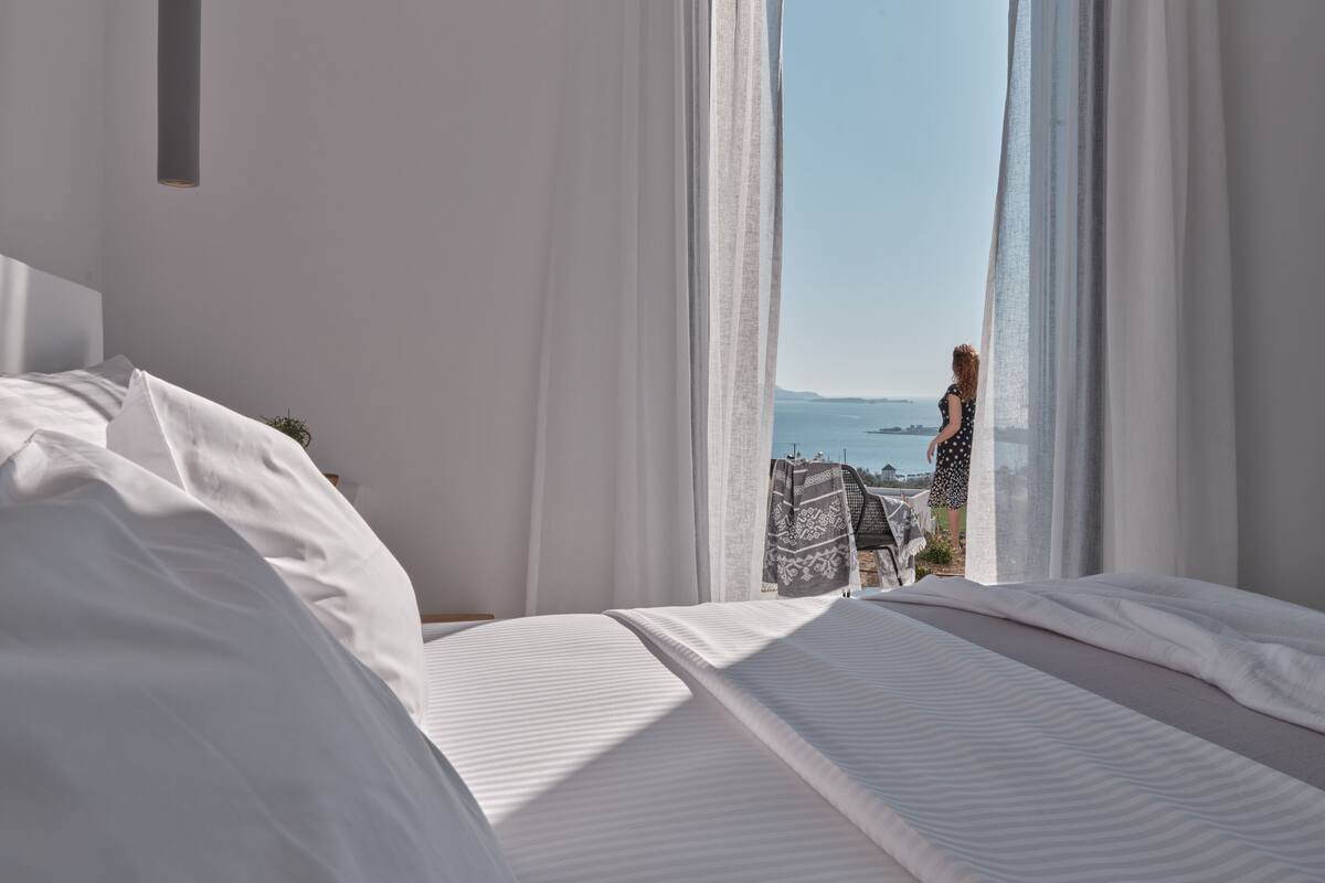 Un dormitorio con una cama grande, ropa de cama blanca y vistas al mar a través de cortinas translúcidas.