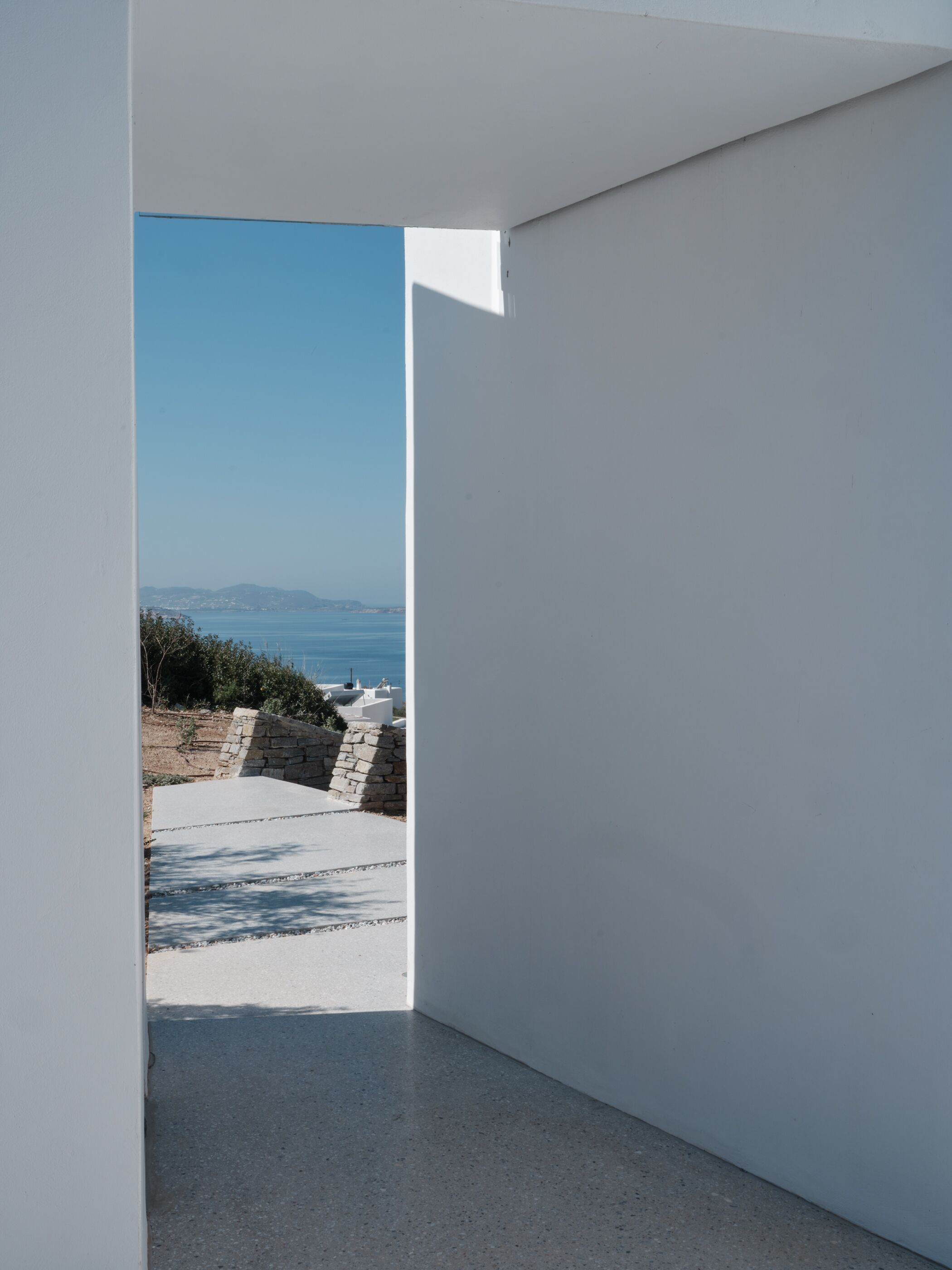 A modern, minimalist room with a clear view of a coastal landscape, including a stone wall and a paved area.