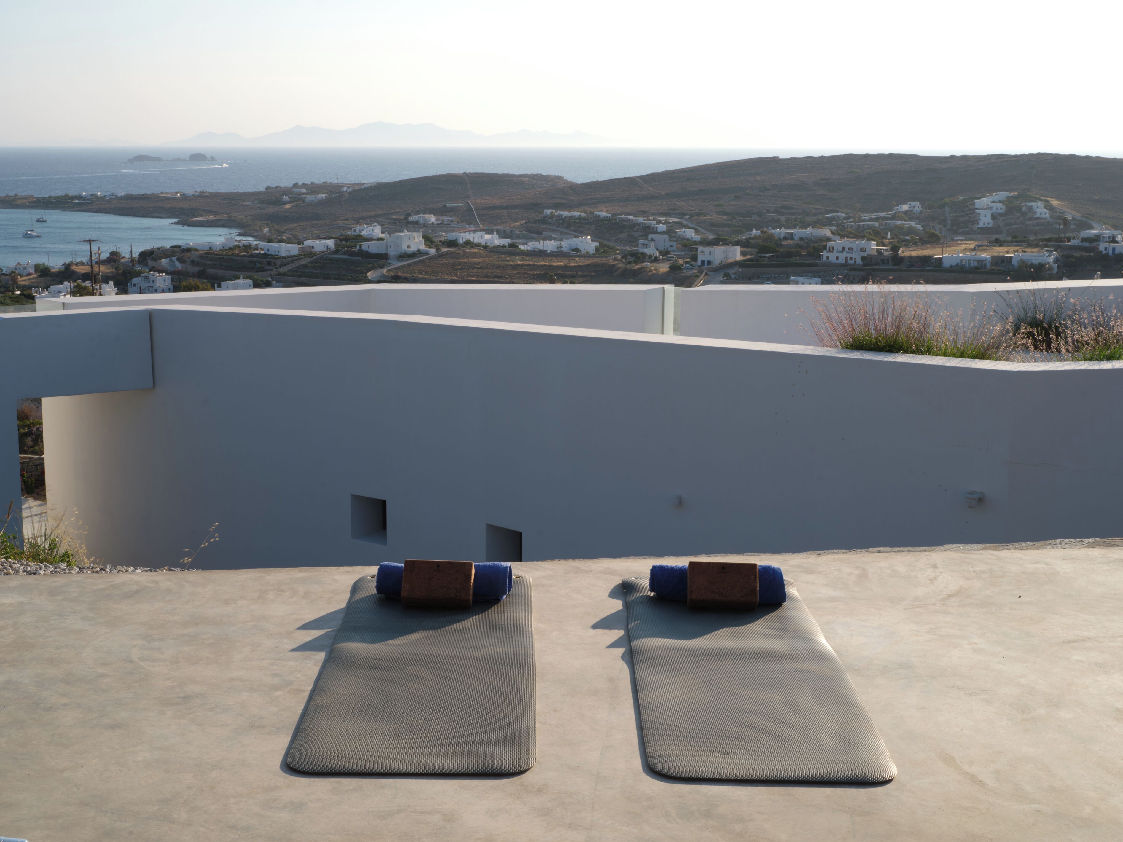 Een terras op het dak, met twee yogamatten en een houten blok, met uitzicht op een kustplaats met witte gebouwen en de zee.