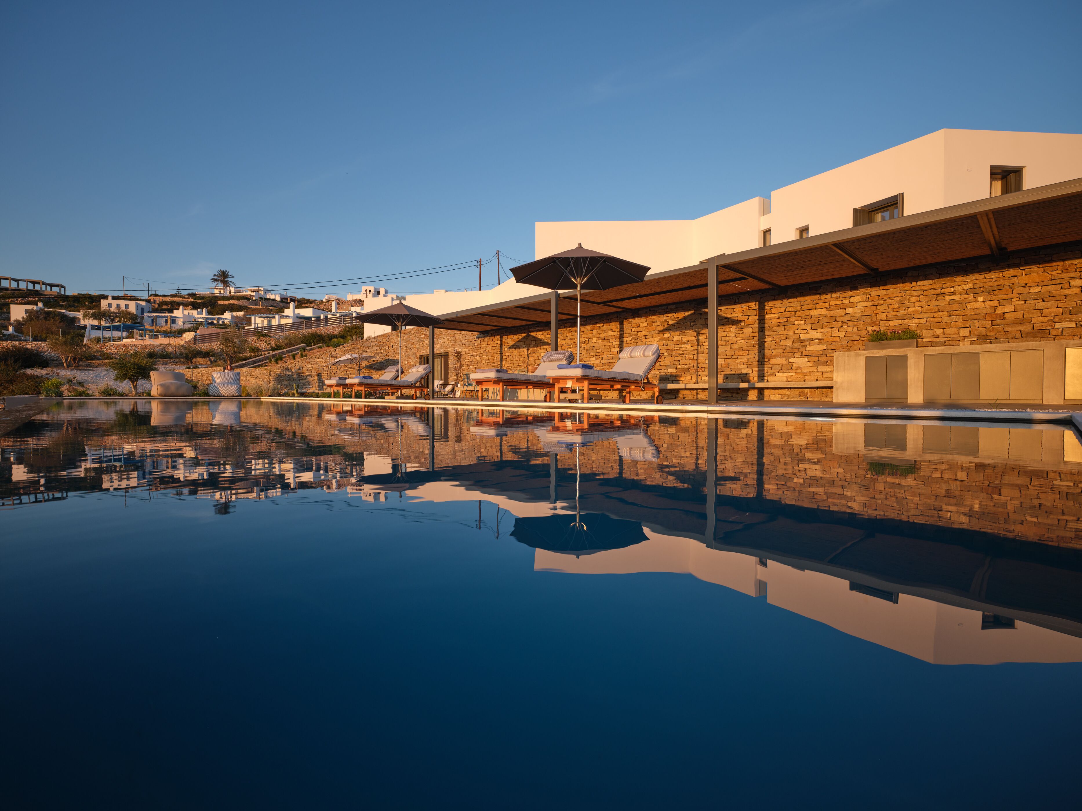 A swimming pool with lounge chairs and umbrellas, surrounded by a stone wall and modern buildings.