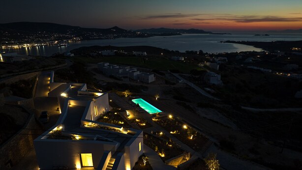 An aerial view of a modern resort with a swimming pool, well-lit pathways, and a view of the coastline at dusk. - Olithos Villas (Paros)