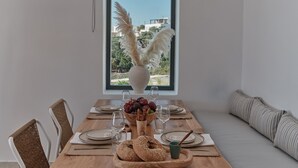 A dining area with a wooden table set for four, a vase with pampas grass, and a view of buildings through a large window. - Olithos Villas (Paros)