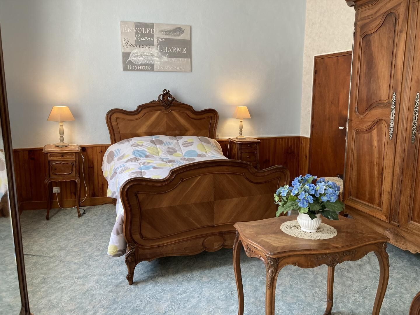 Un dormitorio con cama de madera, mesitas de noche con lámparas, un armario de madera y una pared con un texto enmarcado.